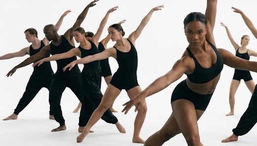 Eine Gruppe Tänzer:innen in Schwarz nimmt dieselbe weit ausgreifende Pose in einem weißen Studio ein, die vorderste Tänzerin blickt direkt in die Kamera.